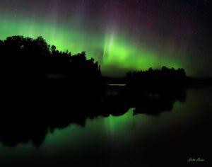 Northern lights illuminating the night sky with green and purple hues reflected over a calm lake and silhouetted treeline at Manitou Weather Station Lodge.